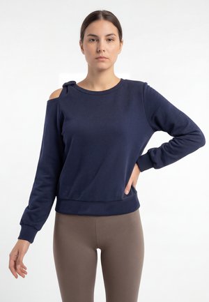 Frau trägt ein marineblaues Langarmoberteil mit einer schulterfreien Aussparung, die mit einem Knoten gebunden ist, kombiniert mit braunen eng anliegenden Leggings, steht vor weißem Hintergrund.