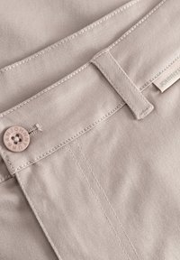 Béžová bavlnená tkanina s hladkou štruktúrou, so šitým pásom, ružovým gombíkom a štítkom na boku.