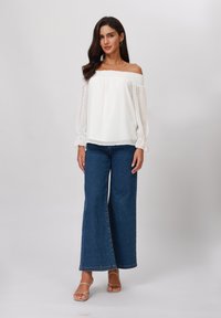 White off-the-shoulder blouse with sheer polka dot texture, puffed sleeves, paired with wide-leg blue denim jeans and beige sandals.