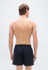 Μαύρα μαγιό σορτς με ελαστική μέση, με λογότυπο στο πλάι. Το ύφασμα φαίνεται λείο και ελαφρύ. Το μοντέλο φαίνεται από πίσω.