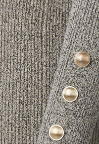 Pull en tricot gris avec des éclats métalliques. Présente une texture côtelée et trois grands boutons ronds avec une finition brillante le long de la manche.