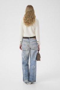 Bílý svetr s texturou, zdobený knoflíky, a roztrhané modré džínové kalhoty. V pravé ruce je držena malá hnědá kabelka.