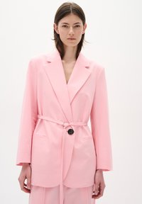 Gaiši rozā džaketis ar brīvu piegriezumu, plašiem klājējiem, jostā siktu vidukli un vienu pogu aizdari. Gluda auduma izpildījums ar divām priekšējām kabatām.