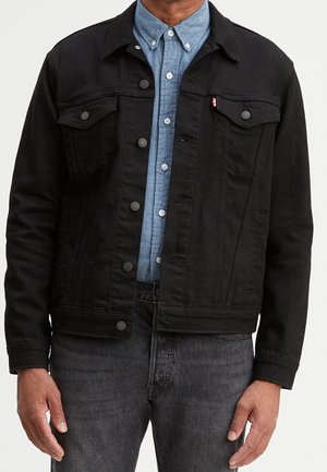Homme portant une veste en jean noire sur une chemise bleue boutonnée et un jean gris foncé, debout les bras détendus le long du corps.