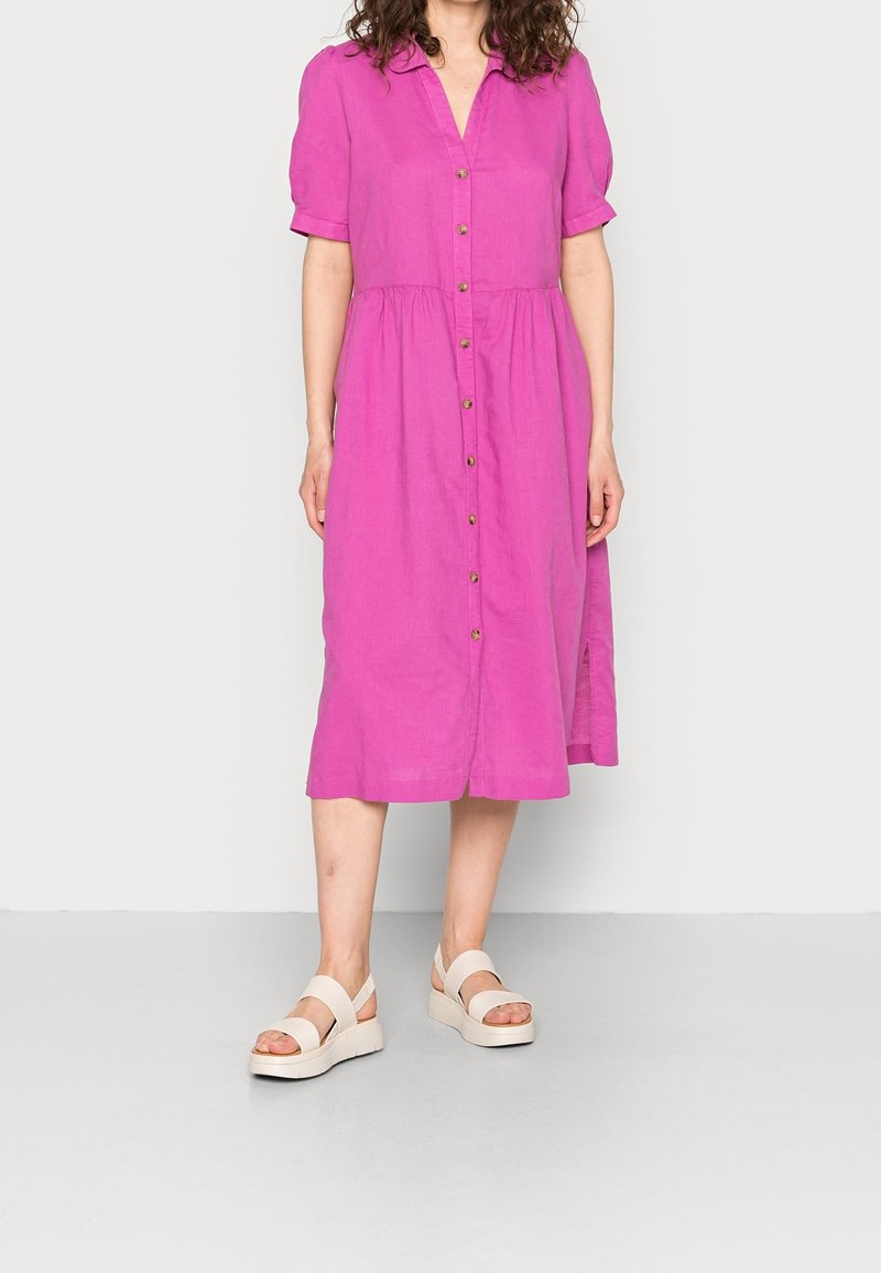 Femme portant une robe midi rose vif avec des boutons sur le devant et des sandales à plateforme beige, debout devant un fond uni.