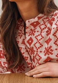 Červená a bílá blůza se vzorem, má mandarínský límec a obsahuje květinové a geometrické motivy. Knoflíky mají hladký, lesklý povrch.