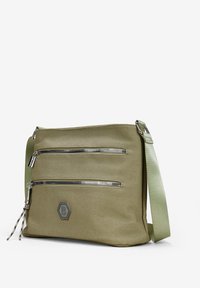 Bolsa de mensajero verde de lona con dos bolsillos con cremallera plateados, un acento de logo hexagonal y una correa ajustable. Textura suave y forma estructurada.
