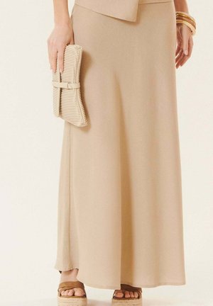 Femme portant une jupe maxi beige, tenant une pochette beige texturée et portant des sandales marron à bouts ouverts avec des bracelets dorés à un poignet.