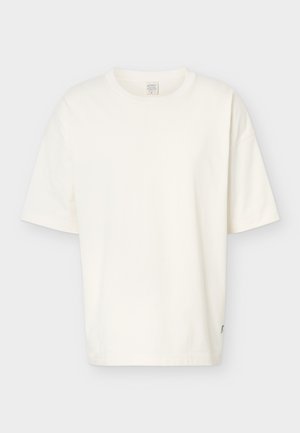 Persoon met kort blond haar die een wit oversized t-shirt draagt met de tekst "Run in Berlin" en een groene teddybeerafbeelding op de rug.