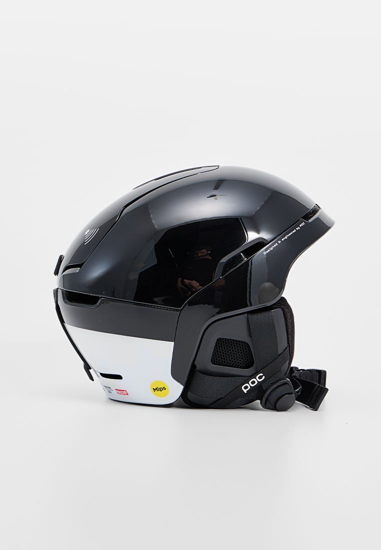 Czarny i biały kask narciarski o gładkim wykończeniu, etykieta technologii MIPS, otwory wentylacyjne, regulowany pasek i pokrętło regulacji z tyłu.
