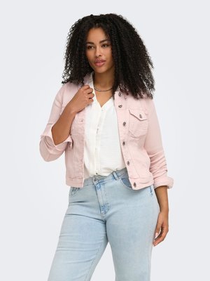 Donna con capelli ricci che indossa una giacca rosa chiaro sopra una camicetta bianca e jeans azzurri, in piedi con una mano vicino al colletto.