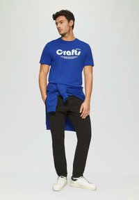 Blauwe katoenen t-shirt met witte tekst, zwarte slim-fit broek en witte sneakers met een marineblauw accent. Sweatshirt geknoopt om de taille.