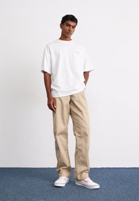 Oversized hvid bomulds T-shirt med blå grafik, beige bukser og hvide sneakers med lyserøde accenter, står på blå tæppe.
