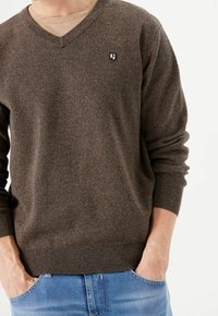 Pull marron en V en tricot texturé. Présente des poignets et un ourlet côtelés, avec un petit logo sur la poitrine. Porté par-dessus une chemise de couleur claire.