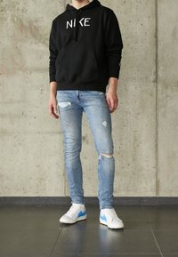 Sudadera negra con el logo de 'NIKE', vaqueros celestes desgarrados y zapatillas blancas con detalles azules, sobre un fondo de hormigón texturizado.