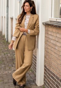 Beige blazer en wijdvallende broek van een lichtgewicht stof, gecombineerd met een witte kanten top. Bruine schoenen met versieringen maken de outfit compleet.