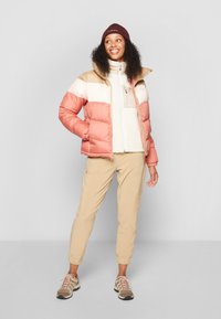 Puffjacka i rosa, beige och krämfärgad, med dragkedja och plyschtextur, i kombination med ljusbeiga byxor och bruna sneakers.