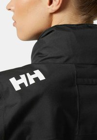 Chaqueta impermeable negra con un alto cuello, textura suave y un logotipo blanco prominente en el hombro. Diseñada funcionalmente con bolsillos.