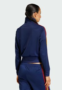 Jakna trenirke mornarsko plave boje s visokim ovratnikom, ukrašena crvenim i žutim prugama na rukavima i uskim pojasom u struku. Glatka tekstura.