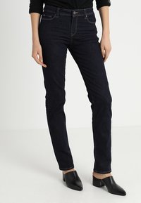 Jeans skinny azul oscuro de denim, con cinco bolsillos, trabillas para cinturón y cremallera. Llevados con zuecos negros.