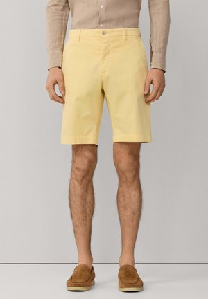 Mann, der hellgelbe knielange Shorts, ein beige langärmeliges Hemd mit Knöpfen und braune Wildleder-Loafers trägt, steht vor einfachem Hintergrund.