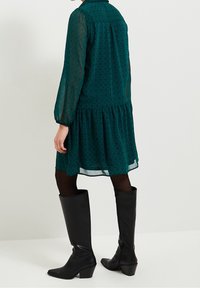 Green patterned dress with long sleeves, tiered hem, and sheer texture. Paired with high black leather boots and dark tights.