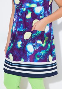 Kolorowa tunika z sukienką o purpurowej bazie z niebieskimi i zielonymi abstrakcyjnymi wzorami, w paskowanej krawędzi i z przednią kieszenią. W zestawie z zielonymi legginsami.