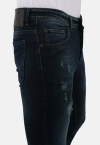 Donkerblauwe denim jeans met een slanke pasvorm, beschadigde details, subtiele vervaging en koperkleurige hardware. Achterpatch met het label "Local Fanatic."