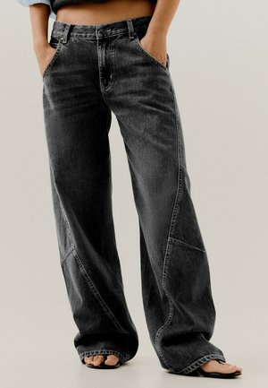 Personne portant un jean en denim noir ample et des sandales noires ouvertes à l'avant, avec les mains dans les poches et un top court visible.