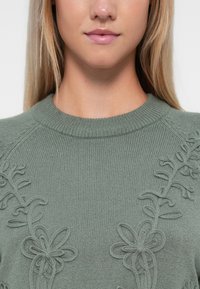 Pull en tricot vert avec un col rond, présentant des broderies florales texturées sur un côté. Tissu doux et texturé avec une coupe confortable.