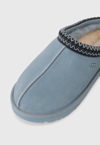 Chausson en daim bleu clair à enfiler avec un liseré cousu noir et blanc et une doublure douce en polaire couleur crème, marqué du logo UGG.