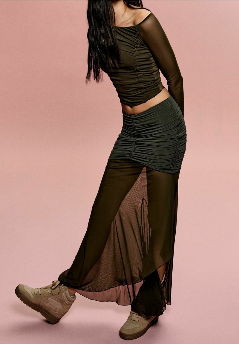 Olivově zelená průsvitná vrstvená halenka a sukně s řasenou texturou. Kombinováno s béžovými vysokými teniskami na jemně růžovém pozadí.