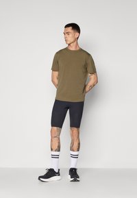 Olivgrünes kurzärmeliges T-Shirt kombiniert mit marineblauen Kompressionsshorts, weißen Socken mit schwarzen Streifen und schwarzen Sportschuhen.