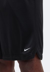Shorts de sport noirs avec un swoosh Nike blanc sur le bas de la jambe droite, portés par une personne sur un fond uni.