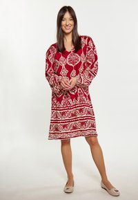 Rochie roșie cu modele florale și geometrice albe, croi lejer, mâneci lungi și decolteu în V. Purtați cu balerini bej. Design simplu.