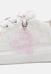 Hvit lær sneakers med gull prikkemønster, dekorert med flettede pastell-lisser, en metallisk tag, og en rosa gjennomsiktig charm festet.