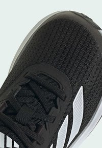 Scarpa da ginnastica nera con un upper testurizzato e sezioni in rete, lacci piatti e tre strisce bianche di branding sui lati.