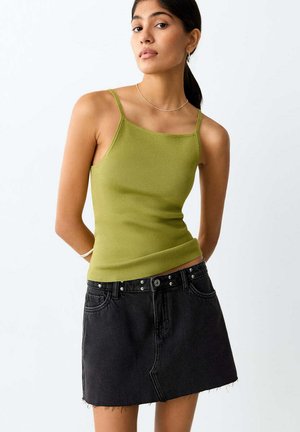 Kobieta ubrana w zielony top bez rękawów z kwadratowym dekoltem i czarną mini spódniczkę jeansową z nitowanymi detalami paska, stojąca z rękami za plecami.