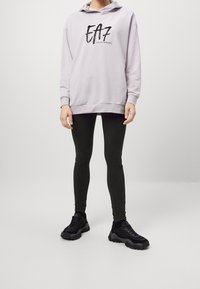 Felpa leggera viola con logo EA7 nero, abbinata a leggings neri e sneakers nere chunky, in piedi contro uno sfondo neutro.