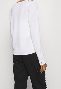 Pull blanc en tricot avec un motif texturé, coupe légèrement ample et détails boutonnés sur les manches, associé à un pantalon noir déchiré.