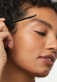 Applicateur de gel à sourcils noir avec une brosse, appliqué sur un teint lisse et bronzé avec une texture de peau visible et une couleur naturelle des lèvres.