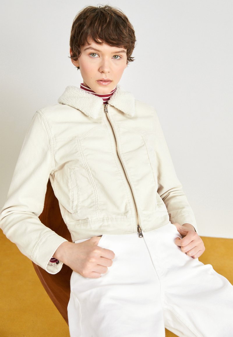 Veste cropped couleur crème avec une fermeture éclair et un col en mouton doux. Présente des poches latérales et un tissu texturé. Associée à un pantalon blanc.