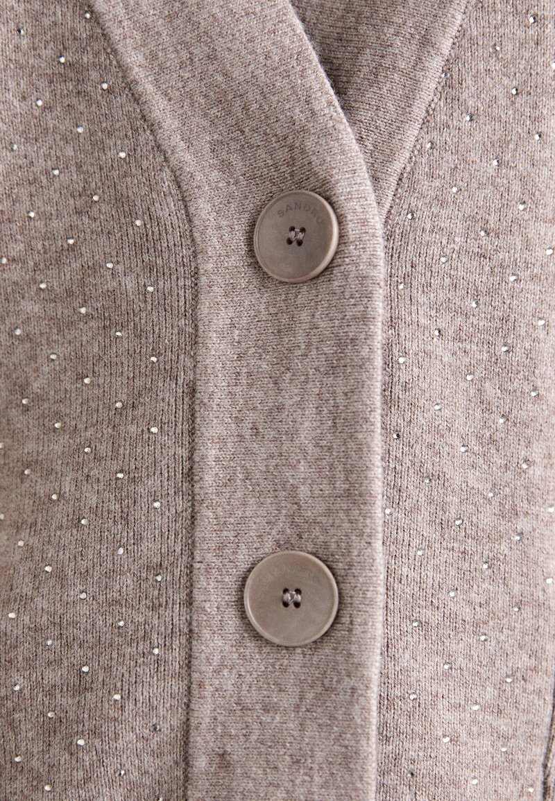 Hellbrauner Cardigan aus strukturiertem Strickstoff, der mit kleinen silbernen Verzierungen und zwei runden braunen Knöpfen auf der Vorderseite ausgestattet ist.