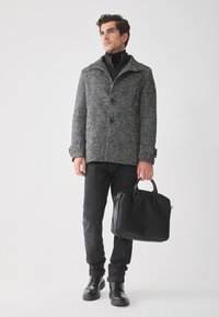 Hombre vestido con un abrigo de espiga gris, pantalones negros y botas negras, sosteniendo un maletín de cuero negro frente a un fondo blanco liso.