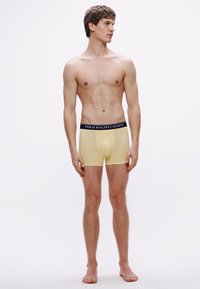 Žlté boxerky s tmavým pásom, na ktorom je logo „POLO RALPH LAUREN“; model stojí vzpriamene, bosý, na bielom pozadí.