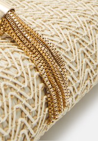 Gros plan sur une pochette tissée beige avec un détail de tassel en chaîne dorée complexe sur un fond blanc.