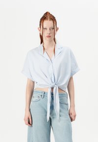 Camisa de botão de manga curta, listrada em azul e branco, com detalhe frontal em nó; combinada com jeans de cintura alta de tom azul claro.
