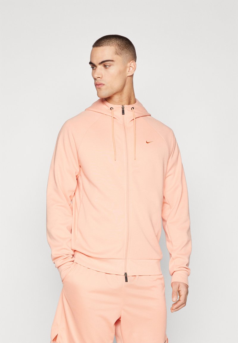 Lyserosa zip-up hettegenser med snorlås, med raglanermer og en liten oransje logo på brystet. Myk stoff og athletic fit.