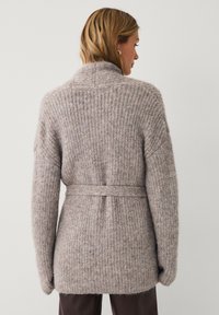 Frau, die einen gerippte, taillierte, langärmlige taupefarbene Pullover mit einem hohen Kragen trägt, von hinten vor einem schlichten Hintergrund gezeigt.