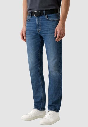 Uomo che indossa jeans blu a gamba dritta, cintura nera, camicia scura e sneakers bianche, in piedi contro uno sfondo semplice.